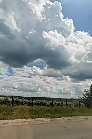 The video shows a scenic view of a rural landscape under a cloudy sky. The foreground features a paved road with a gravel shoulder. Beyond the road, there is a grassy embankment with various trees and bushes. In the background, rolling hills are visible, dotted with what appear to be vineyards or fields. Large, puffy white clouds dominate the sky, with patches of blue sky peeking through. The sun is shining, casting a bright light on the scene. The overall impression is one of a peaceful and picturesque countryside on a partly cloudy day.