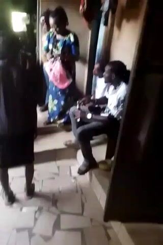 The video shows a group of people inside what appears to be a modest home.  A woman in a floral dress holds a pink bag and speaks to someone out of frame near the doorway.  Another woman stands next to her wearing a tan dress and sunglasses.  Two men are seated on a step, one is wearing a white shirt and jeans, and appears to be drinking from a bottle. Another child is squatting on the floor. A younger child wearing a blue sleeveless shirt and red shorts sits on the floor. The interior of the home is simple, with tiled floors and painted walls.  Some personal items hang on the walls.