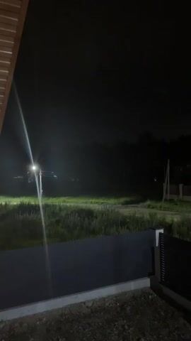 The video shows a night scene from a covered porch. A bright light emanates from the left side of the frame, casting a strong glare across a grassy field. The field is somewhat overgrown, with tall grass and some shorter vegetation. In the distance, silhouetted against the night sky and illuminated by the bright light, are what appear to be the shapes of trees or a forest line. A dark fence runs horizontally across the middle ground of the image, separating the foreground from the field. A light pole with a fixture is visible in the field, serving as the source of the bright light. To the right of the field, a wooden utility pole with wires is visible. The foreground shows the edge of a concrete foundation and loose gravel. The overall atmosphere is dark, with the primary light source creating a hazy, illuminated effect in the distance.