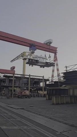 A large gantry crane with a red horizontal beam and white striped vertical supports is operating in an outdoor industrial setting. The crane is lifting a cylindrical metal structure, which appears to be part of a larger construction project. Below the crane, there are other construction elements, including scaffolding, metal beams, and what looks like a forklift. The ground is covered in gravel and there are train tracks in the foreground. The sky is overcast.