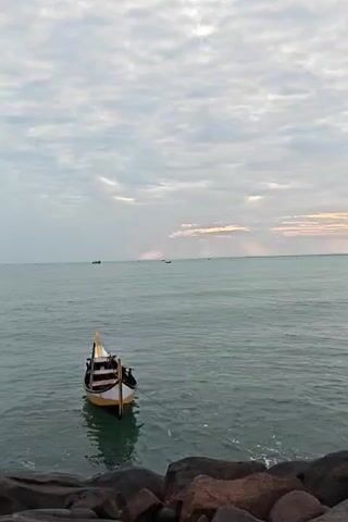 The video shows a stationary boat tied to the rocky shore of an ocean. The boat is mostly yellow and white and sits calmly on the water, which appears flat with gentle ripples. A rope tethers the boat to the dark grey rocks at the shoreline. In the distance, two more boats are faintly visible on the water's surface. The sky overhead is a mix of light grey and white clouds, with streaks of sunlight breaking through the cloud cover on the horizon. The general atmosphere suggests a calm and somewhat overcast day.