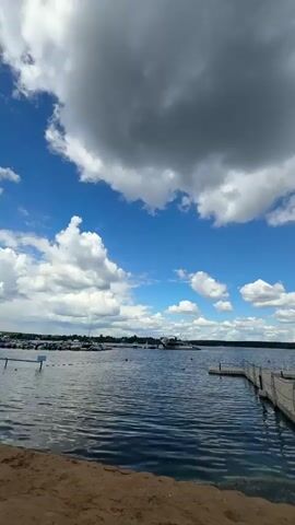This is a vertical video showcasing a serene lake scene under a dramatic sky. The foreground is a sandy beach, leading to a tranquil lake with ripples on its surface. A long, white modular dock extends from the right side of the frame into the water, with a smaller, detached section of the dock further out.

In the middle ground, across the lake, a distant shoreline is visible, dotted with trees. There's a small cluster of boats moored in the distance on the left side of the frame, suggesting a marina or a place for recreational boating. The water itself appears clear and calm, reflecting the sky.

The sky is the most striking element, a vast expanse of vibrant blue punctuated by large, fluffy white and dark grey clouds. The contrast between the bright blue sky and the ominous, heavy clouds creates a dynamic and visually interesting atmosphere.

The time of day appears to be daytime, likely late morning or early afternoon, given the bright sunlight filtering through the clouds. The weather seems to be transitioning, with the possibility of rain or a storm approaching, adding a sense of drama to the otherwise peaceful scene.

The overall mood is peaceful yet awe-inspiring, with the power of nature evident in the sky. The absence of people suggests a quiet moment, perhaps early in the day or in a less crowded season. The location, identified as a tourist resort on the Klyazma Reservoir in Russia, hints at a recreational or vacation setting.

There is no visible text in the media file.