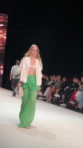 A fashion show is underway. On a white runway, a blonde female model walks towards the camera. She is wearing a white bra, a white jacket, and a long, flowing, sheer green skirt with ruffles. She also has on sunglasses and large silver earrings. She is holding a white clutch purse in her left hand. In the background, to the left, another male model walks away from the camera. He is wearing a striped shirt and dark pants. To the right of the runway, an audience is seated in rows, watching the show. The background is dark.