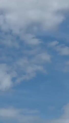 A Ryanair airplane is shown in flight against a blue sky with scattered clouds. The airplane is white with blue and yellow markings. The word "RYANAIR" is visible on the side of the fuselage. The airplane is shown from the side, with its engines visible. The propellers on the engines appear to be spinning. The airplane is moving from left to right in the frame.