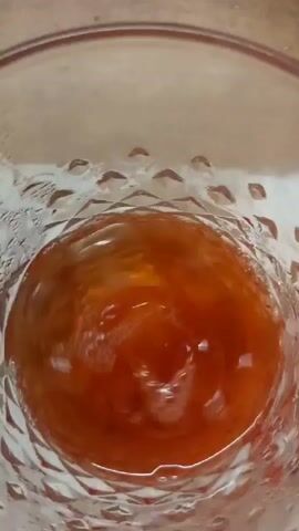 A close-up shot from directly above a glass shows a reddish-orange liquid with ice cubes. The glass has a textured pattern around the rim and appears to be slightly condensation-covered. The liquid is disturbed by the movement of the ice, creating small bubbles and ripples on the surface.
