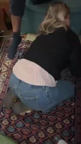 A woman with blonde hair wearing jeans, a black sweater, and a pink and white striped shirt underneath, kneels on a patterned rug. She is cleaning a stain on the light gray couch with a blue cloth. There are two large red Santa boot decorations resting on the couch next to her. A wine bottle lies on the rug behind her. Several people are partially visible near the couch.