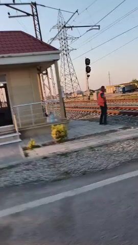 The video is a shaky, first-person perspective of a moving vehicle. The scene is an outdoor, rural environment with a clear sky. On the left, there is a small, light-colored building with a red roof and a window. Next to it is a small tree with red fruit. Further in the background, there are several large metal electrical towers and power lines. On the right, there are trees and bushes, including a large green conical tree and a rounded green bush. The vehicle appears to be driving on a road next to a sidewalk or paved area.
