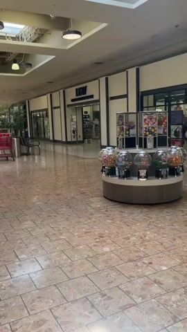 A POV shot moves through a mall hallway. A shopping cart is visible on the left. To the right, a row of gumball machines sits on a circular display. Further down the hall, a store sign reads "DGW". On the left, a store window displays balloons and other items. Two people are walking away from the camera in the distance.