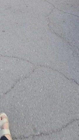 The video begins with a close-up shot of cracked asphalt. A bare foot in a sandal is visible in the lower left corner of the frame, appearing to walk away from the camera. The viewpoint then shifts to follow the person walking. A black car is partially visible on the right side of the frame. The path continues along the asphalt, with some gravelly patches. In the distance, a metal pole is seen fixed to the ground, with white and black striped pipes extending from it. The camera continues to move forward, showing the pole and pipes more clearly. The scene appears to be an outdoor area, possibly a road or parking lot, with a somewhat utilitarian environment.