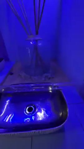 A close-up shot under blue lighting shows water flowing from a clear, horizontal tube into a sink. The sink is a square shape with rounded corners, and a metallic, speckled surface. A black drain is visible in the center. Behind the sink is a large, clear glass vase containing dried reeds. The vase sits on a wooden plank scattered with small white rocks. A small, partially visible white object is situated on the left of the frame.