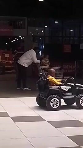 The video shows an indoor scene, likely a shopping mall or large store, with a tiled floor in a black and white checkerboard pattern. In the foreground, a young child is seated in a black and white electric toy jeep. A man in a white soccer jersey, blue jeans, and black boots is walking towards the toy car, seemingly interacting with the child. In the background, another man is leaning over a shopping cart where a child is sitting. There are other people visible in the background, as well as a sign that reads "TRY/ KUINCIA".  The lighting is bright but slightly dim, typical of indoor shopping areas.