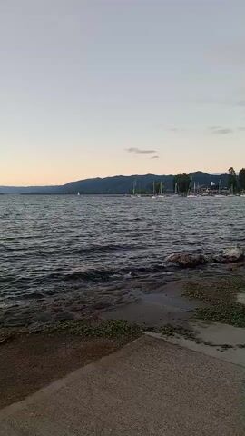 The video captures a panoramic view of a large lake or reservoir, likely Lake San Roque in Villa Carlos Paz, Argentina, during sunset or dusk. The sky transitions from a soft light blue overhead to warm orange and pink hues along the distant horizon, where mountains and hills are silhouetted.

The camera pans slowly from left to right, revealing the expansive body of water with visible waves and ripples, suggesting a light breeze. In the distance, several sailboats are present; some appear to be docked along a distant shore or in a marina on the right side of the frame, while one small sailboat sails in the open water.

In the foreground, a rocky and concrete shoreline meets a paved path or promenade. A stone marker with an arrow pointing left is visible at the 0:01 mark. As the camera continues to pan, a lamppost and the partial silhouette of a person standing near the water's edge can be seen at the 0:03 mark, observing the serene evening landscape.