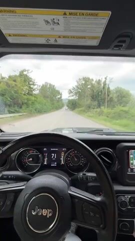 This video shows the view from the driver's seat of a Jeep, looking forward through the windshield. The Jeep is on a paved road with trees and foliage lining both sides. The dashboard displays the speed as 70 km/h, and the temperature is 20°C. The sky is overcast. There are other vehicles visible in the distance on the road. The sun visors are down and contain warning labels. The driver's hands are not clearly visible, but implied by the steering wheel.