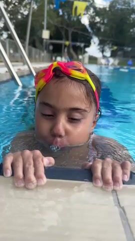 A young girl wearing pink and yellow swim goggles is in a swimming pool. Her hands are gripping the edge of the pool. She exhales bubbles into the water. The camera is positioned close to her face, at eye level. The background shows a swimming pool area with trees and flags.