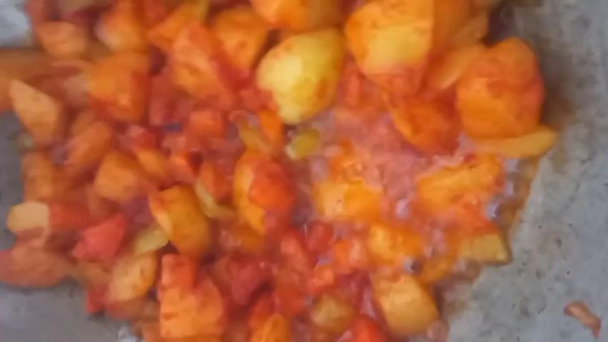 This close-up, top-down shot showcases a batch of diced potatoes being cooked in a pan. The potatoes are cut into irregular, bite-sized pieces and are mixed with a vibrant red sauce, possibly tomato-based, and some green ingredients that appear to be sliced bell peppers or chilies. The dish is sizzling and steaming, indicating it is actively being prepared. The cooking vessel appears to be a gray, non-stick pan with a textured surface. The lighting suggests it might be daytime, but the specific time of day and weather conditions are not discernible from the image. No people or text are visible in the frame. The focus is entirely on the food being cooked, capturing the textures and colors of the ingredients in the process of becoming a meal. The scene is likely a kitchen setting in Ынтымак, Kyrgyzstan, given the provided location context.
