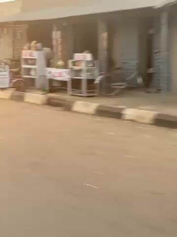 The video shows a street scene in what appears to be an African town or city. The road is paved and has a curb painted with alternating black and white stripes. On the right side of the road, there are several small shops or stalls. People are seen walking and standing around. Several motorcycles are also visible. One person is seen riding a motorcycle down the street. A black car drives past the camera. The sky is a pale orange color, suggesting it is either early morning or late afternoon. The overall impression is that of a busy, everyday scene in a developing area.