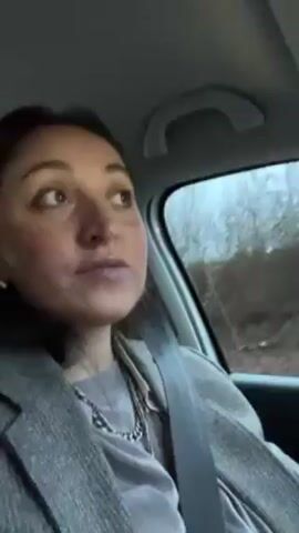 The video shows a woman in a car, facing the camera and pouting her lips. She holds up a glass jar with a lid and a brown sleeve. The perspective shifts to show the view from the driver's seat as the car moves along a wet road. Other cars are visible ahead, and trees and a sound barrier are seen on the side of the road. The sky is overcast.