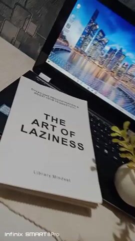 A book titled "The Art of Laziness" is displayed on top of a laptop. The book cover is white with black text. The laptop screen displays a city skyline. A small potted plant is visible on the right side of the frame. The scene is illuminated by natural light.