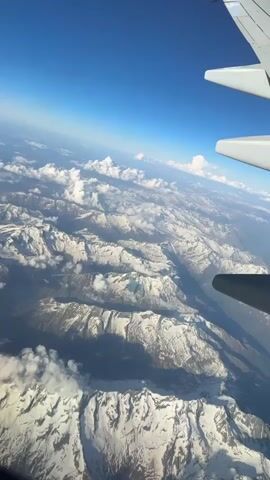 The video shows an aerial view of snow-covered mountains from an airplane window on a clear, sunny day. The airplane's wing is visible in the upper right corner of the frame. The mountains are jagged and covered in patches of snow, with some clouds hovering around the peaks. The sky is a deep blue.