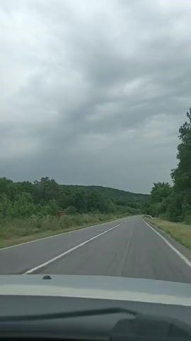 The video shows a first-person view from a vehicle traveling on a two-lane asphalt road. The road is surrounded by lush green trees and vegetation on both sides, with rolling hills in the distance. The sky is overcast with gray clouds, suggesting a cloudy or potentially rainy day. The road has white lane markings. There are no other vehicles or people visible in the immediate vicinity. The camera appears to be mounted on the dashboard or windshield of the vehicle.