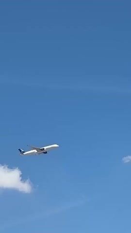 A wide shot captures an airplane in flight against a bright, clear blue sky. The airplane, predominantly white with blue accents on its tail, is positioned in the upper right quadrant of the frame. It appears to be in ascent, leaving a faint, wispy contrail behind it that stretches diagonally across the sky. A small, fluffy white cloud is visible in the lower left corner of the image. The lighting suggests it is daytime, likely midday given the brightness and the absence of long shadows. The scene is serene and peaceful, evoking a sense of travel and open skies. There is no visible text in the media.