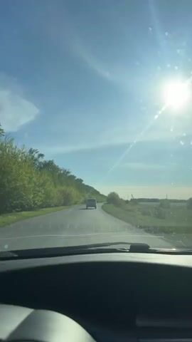 The camera is inside a car, driving down a road on a sunny day.  The sky is blue with wispy clouds, and the sun is bright, creating glare on the windshield.  There's another car ahead on the road. The roadside is lined with green trees and fields. The car dashboard is visible in the foreground. Music is playing.