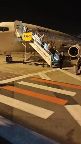At night, passengers disembark from a Hebei Airlines airplane using a mobile staircase. Several individuals, including a woman in a dark coat and a face mask pulling a suitcase, descend the stairs and walk across the striped pedestrian crossing on the tarmac. There is also a woman in a red sweater with a little girl walking alongside. The airplane is brightly lit, with the airline's logo visible. The scene takes place on an airport apron, with other aircraft visible in the background.