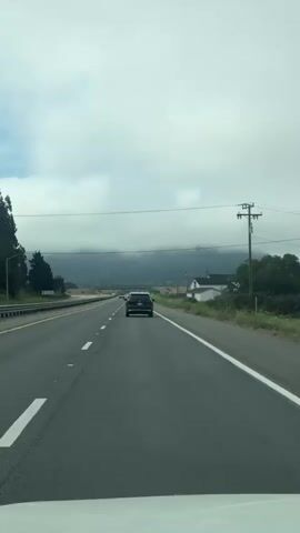 The video shows a first-person perspective of driving on a highway. The road has multiple lanes and is marked with white lines. Ahead, a black SUV is driving in the same direction. On the right side of the road, there are trees and a white house with a black roof. In the distance, a mountain range is visible, partially obscured by clouds. The sky is overcast with dense clouds. The camera is positioned low, possibly from a dashboard, as indicated by the white bumper visible at the bottom of the frame.