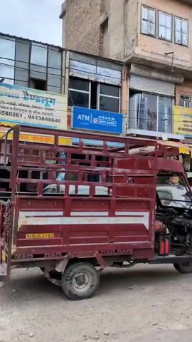 The video shows a street scene, likely in India, with various vehicles and buildings. In the foreground, a white Toyota Etios Cross is parked on the left, followed by a silver Chevrolet Enjoy MPV. A green auto-rickshaw is visible on the far right, partially obscuring the view. The background features a row of shops and businesses with signs in Hindi. One sign reads "Shri Gahlot Henpalum." Further down the street, a white Isuzu D-Max pickup truck is parked. Several people are walking on the sidewalk, including a woman in a traditional Indian dress and two men. Another scene shows a truck with a cage-like structure on its back, a white Isuzu pickup truck, and a person in a yellow shirt on a motorcycle. The buildings have intricate architectural details, characteristic of older structures. The street appears to be paved with asphalt. The overall impression is a bustling street in an Indian city.