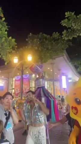 The video shows an outdoor performance at night. The street is lit by lampposts and the fronts of shops. A person on stilts is dressed in a colorful skirt and a headpiece.  A person on the ground is wearing a shiny silver vest. A large sun-shaped cartoon character is visible on the right. The silver vest wearer waves to the camera while smiling. Other people are visible watching the performers.
