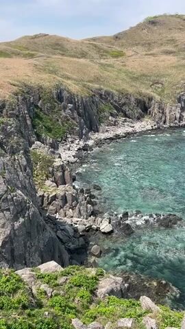 The video shows a high-angle shot of a rocky coastline. The water is a clear turquoise color near the shore, becoming a darker blue further out. The coastline is composed of jagged, gray cliffs and rocks. There is a hill in the background covered with dry grass. At the top of the hill, a small white building with a red roof is visible. The sky is overcast and appears to be a light gray color. The camera pans slowly to the right, revealing more of the coastline. There are no people visible in the video. The focus of the shot is on the natural landscape, highlighting the contrast between the rugged cliffs and the clear water.