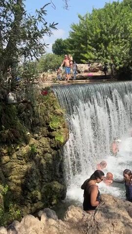 The video shows a group of people enjoying a natural waterfall and swimming area on a sunny day. In the foreground, several individuals, including women and children, are wading in the turquoise water near a rocky edge. One young girl in a swimsuit is reaching down towards the rocks. Behind them, a cascade of water falls from a man-made structure, creating a refreshing spray. A man stands on the edge of the waterfall, arms outstretched, possibly preparing to jump. Other people are seen swimming and bathing in the pool beneath the falls. Lush green trees and foliage surround the area, suggesting a natural park or recreational spot. The water is clear and inviting, with some people using snorkeling gear. The overall atmosphere is one of leisure and outdoor recreation.