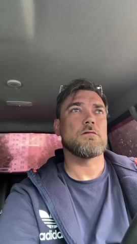 The media file is a close-up video of a man sitting inside a vehicle. He has short brown hair, a beard, and is looking slightly upwards and to his left. He is wearing a dark blue t-shirt with the Adidas logo visible on the left chest, and a dark blue zip-up hoodie over it, also with the Adidas logo on the left chest. Sunglasses are perched on top of his head.

The background visible through the car windows shows a building with pinkish-red tinted windows and trees. The interior of the car is visible, including the grey headliner with a light fixture and a red strap.

The man appears to be speaking to the camera, though the audio is not provided. His expression is neutral, with a slight furrow in his brow. The lighting suggests it is daytime. The provided location context places this scene in Usinsk, Russia.