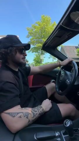 A man with a beard and a large tattoo on his left arm drives a convertible car on a sunny day. He is wearing a black t-shirt, black shorts, sunglasses, and a black baseball cap. He has gold bracelets on both wrists. The car has red leather seats. The top of the car is down. The man is looking to his right as he drives. The car is moving through a neighborhood with houses and trees visible in the background. Some houses have solar panels on the roof. There are also colorful flags strung across the street.
