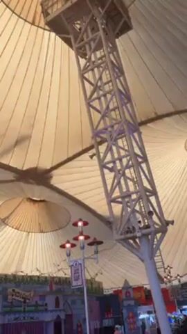 The video shows an indoor space with a large white fabric canopy supported by a tall, slim, tower-like structure made of crisscrossing white beams. The canopy is wrinkled, and there are circular holes at the top, presumably for ventilation or light. The tower structure is in the center of the canopy and reaches almost to the top. At the base of the tower are signs and decorative objects. The lower part of the video shows people walking around, tables, and a few structures that look like part of an indoor attraction. The camera moves and pans upwards.