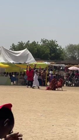 The video shows a gathering of people in an open, sandy area, seemingly for a cultural event or ceremony. There are several individuals dressed in traditional attire, including two figures in the center of the frame: one in a red robe and another in a white robe. They seem to be performing or interacting with a person seated on a chair, draped with a red cloth.  The audience is seated under tents or standing around with umbrellas, observing the event. In the background, there are trees and buildings, as well as a communication tower. The overall atmosphere suggests a public celebration or cultural performance.