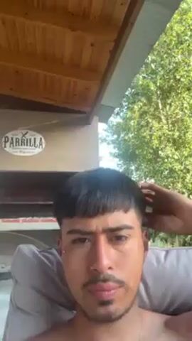 The video features a close-up of a young man with dark hair and a short, blunt fringe (bowl cut). He has a nose ring and a small stud earring. He is sitting in what appears to be a grey cushioned chair, possibly outdoors. Behind him is a wooden ceiling or overhang, part of an outdoor structure. A sign that reads "PARRILLA" is visible in the background, along with a brick structure possibly a barbecue or grill area.  Outside the structure, green foliage is seen. The man is touching his hair and looking directly at the camera. The overall lighting suggests it is daytime.