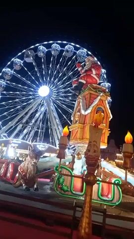 At night, the camera pans across a Christmas market scene. The focus starts on a lit-up carousel ride with Santa Claus on top of a house-like structure. A Ferris wheel glows in the background. As the camera pans, the scene reveals people walking across a red carpet. A large inflatable snowman stands next to a candy cane decoration. The background shows a row of buildings with distinctive gabled roofs. People are milling around, enjoying the market atmosphere.