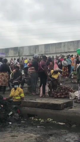 The video depicts a bustling outdoor market scene. Many people are present, some carrying goods on their heads. The ground appears to be uneven and littered with debris. In the background, there is a large concrete wall and some structures that look like market stalls. A poster with a person's image is visible in the distance. Several vehicles are also present, including a white van and a maroon SUV. Some individuals are seen interacting with each other, while others are engaged in their activities within the market. The overall atmosphere suggests a lively commercial environment, albeit one with significant environmental challenges, indicated by the visible waste and unkempt ground.