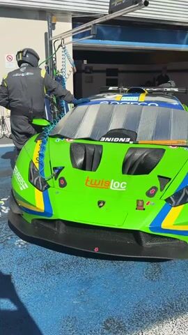 The video shows a green Lamborghini race car being serviced by a team of mechanics in a garage. The car is brightly colored with blue and yellow stripes. One mechanic is working on the front of the car while others are nearby. The garage is well-lit and has a blue floor. The camera then pans out to show a line of race cars, including the green Lamborghini, lined up on a race track. The mechanics are standing near the cars, and people are milling about in the background. The sky is clear and blue.