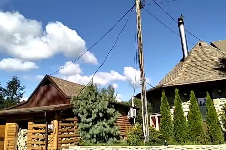 The video shows a rural outdoor scene with several structures and natural elements. In the foreground, there are two parked vehicles: a black car and a blue van. Behind them, a wooden building is visible, with its facade constructed from logs and a corrugated metal roof. To the left of this building, a stack of firewood is neatly arranged against the wall. Further back, a taller, dark-colored metal structure, possibly a tower or antenna, stands against a backdrop of dense green forest. The sky is blue with scattered white clouds. Another building with a similarly pitched, dark-colored roof is visible on the right side of the frame. The overall impression is one of a rustic, possibly mountainous, environment.