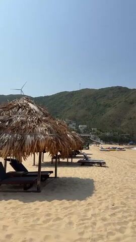 The video shows a beach on a sunny day, panning right to show the ocean. In the foreground, there are thatched umbrellas with lounge chairs beneath them on the sand. A green, mountainous hill rises in the background behind the beach. At the foot of the hill, there are several buildings. Further along the beach, closer to the ocean, there are a few people and some kayaks. As the camera pans right, the hill becomes a rocky outcropping on the right side of the frame. The ocean water is clear and turquoise. The sky is a cloudless blue.