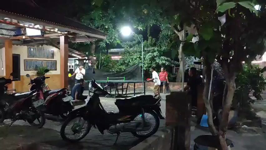 At night, a group of people are playing table tennis in an outdoor area. Several motorcycles are parked in the foreground, with one partially obstructing the view of the table tennis players. A building with a roof is visible on the left, and a large tree with green leaves is on the right. The area is lit by a street lamp. There are at least four people actively playing or watching the game. One person is on the left side of the table, wearing a white shirt and shorts, holding a paddle. Another person is on the right side of the table, wearing a red and white striped shirt and shorts, also holding a paddle. A third person is seated on a low wall or bench on the right, observing the game. A fourth person is partially visible near the building on the left, appearing to be kneeling or crouching. The overall atmosphere is casual and relaxed.