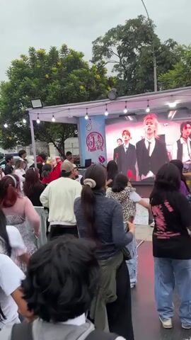 The video shows a group of people dancing to a K-pop song in front of a stage, outdoors. A large screen on the stage displays a K-pop music video. The dancers seem to be following along with the dance moves from the video. The setting appears to be at a festival or outdoor event, with trees visible in the background. There are people of various ages and ethnicities present. The weather looks overcast.