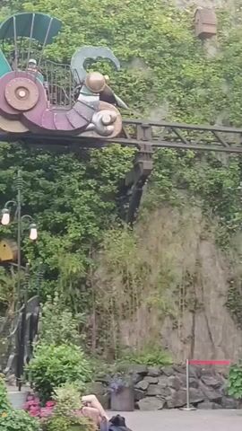 The video pans across a scene with a roller coaster track winding through lush greenery and against a stone wall. Several birdhouses are visible on the wall. At the bottom of the frame, four women wearing hijabs are seated under a simple canopy, with one woman raising her hand. The roller coaster itself appears to be a whimsical, steampunk-inspired vehicle. Plants and flowers are scattered throughout the foreground.