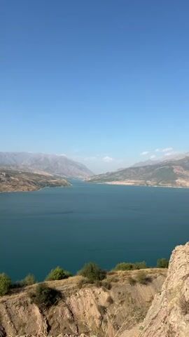 The video shows a sweeping panoramic view of a large, turquoise body of water surrounded by arid, mountainous terrain. The water appears calm, reflecting the clear blue sky. The landscape is characterized by rolling hills and rugged cliffs with sparse vegetation, primarily consisting of dry grasses and scattered shrubs and trees. In the distance, larger mountain ranges are visible, fading into a hazy atmosphere. The viewpoint seems to be from an elevated position on a hillside, overlooking the expanse of water and land. There are no people or artificial structures immediately visible in the foreground or mid-ground of the frame. The overall impression is of a vast, natural landscape under a bright, sunny sky.