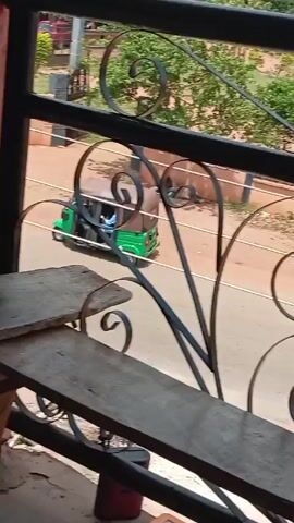 A video shows an elevated view through a black, wrought-iron balcony railing. The foreground contains a wooden shelf, possibly makeshift, sitting atop a support structure. A red-brown textured post stands behind the shelf.

The camera focuses through the railing onto a street below.  A green auto-rickshaw and a yellow auto-rickshaw drive on the road. Later, a motorcycle with three riders passes by.  The environment includes trees, power lines, and buildings visible in the background. The overall setting appears to be a residential or commercial area.