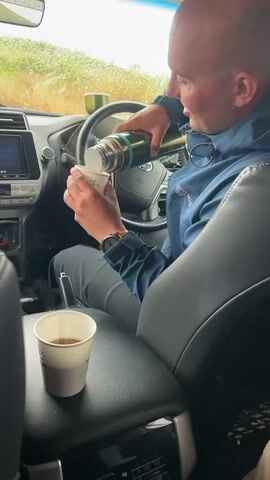 Inside a car, a bald man wearing a blue jacket pours tea from a green and silver thermos into a white paper cup. He sits in the driver's seat and smiles as he finishes pouring. Another person holds a similar paper cup in the foreground, with tea already in it. Outside the car, rain is visible on the windshield. The car's interior is gray, and the dashboard and steering wheel are visible.