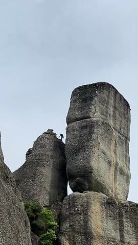 A wide shot captures a tall rock formation against a gray sky. A solitary person is seen atop a smaller rock formation adjacent to the larger one. The larger rock is substantial, featuring a rectangular shape balanced on a broader base. A small patch of greenery is visible on the side of the smaller rock. The scene is still, with no visible action beyond the figure's presence on the rock.
