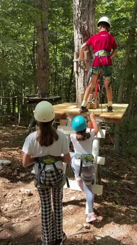 Three children wearing helmets and safety harnesses are in a forest adventure park. Two girls are on the ground near a wooden ladder leading up to a platform built around a tree. A boy is already on the platform, adjusting his safety equipment. In the background, other platforms and rope bridges are visible, suggesting a full course. The ground is covered with leaves and dirt, and tall trees with green foliage surround them. The sunlight filters through the tree canopy, creating dappled light on the forest floor.